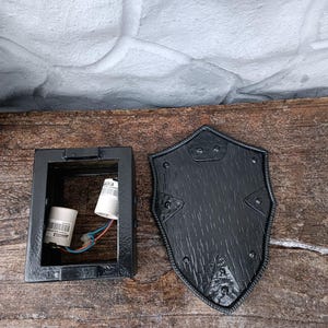 May include: A black metal wall sconce with a shield-shaped cover. The sconce has a rectangular base with a light fixture inside. The shield cover has a textured wood grain appearance and is secured with rivets.