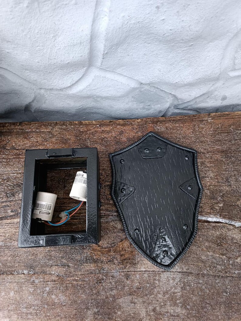 May include: A black metal wall sconce with a shield-shaped cover. The sconce has a rectangular base with a light fixture inside. The shield cover has a textured wood grain appearance and is secured with rivets.