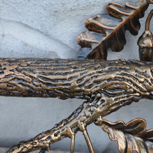 May include: A close-up of a metal sculpture of a tree branch with leaves and acorns. The branch is textured to look like wood and is painted in a gold and brown color.