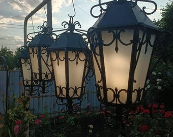 May include: Three black wrought iron lanterns with frosted glass panels hang from a metal frame in front of a garden with green foliage and pink flowers.