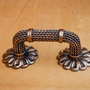Könnte beinhalten: Ein dekorativer Schrankgriff mit einem seilartigen Design und einer blütenförmigen Basis. Der Griff ist aus Metall und hat eine Bronze-Oberfläche.