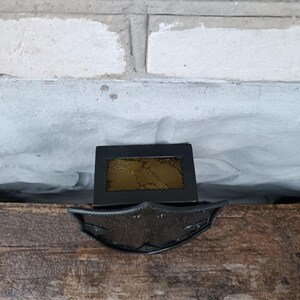 May include: A black metal decorative piece with a rectangular frame and a decorative, shield-like base. The frame holds a gold-colored, textured insert. The piece is set on a wooden surface against a white brick wall.