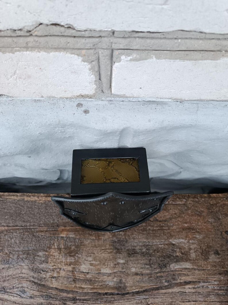 May include: A black metal decorative piece with a rectangular frame and a decorative, shield-like base. The frame holds a gold-colored, textured insert. The piece is set on a wooden surface against a white brick wall.