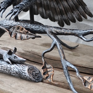 May include: A metal sculpture of an owl perched on a branch with a small mouse on the branch below. The owl is made of black metal and has a detailed wingspan. The branch is made of brown metal and has leaves made of copper metal.