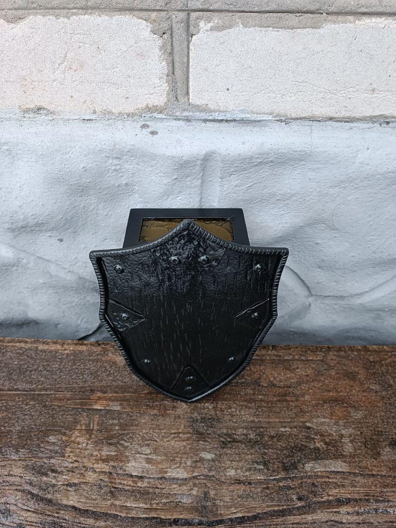 May include: A black, shield-shaped wall sconce with a textured surface and decorative rivets. The sconce is mounted on a wooden surface against a white brick wall. The sconce has a rectangular top with a glass panel.