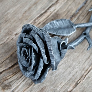 Könnte beinhalten: Eine Metallrose mit grauer Oberfläche. Die Rose ist aus Metall gefertigt und hat ein realistisches Aussehen. Die Rose liegt auf einer Holzoberfläche.