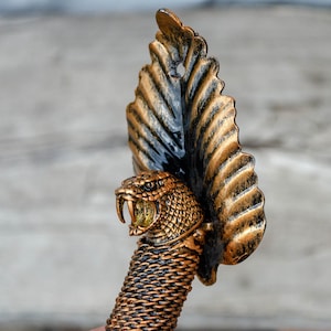 Puede incluir: Un tapón de botella de metal dorado y negro con forma de cobra con la capucha levantada. Los ojos de la cobra son amarillos y sus colmillos son visibles.