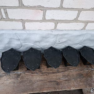 May include: Six black, shield-shaped decorative items with rivet details are arranged on a wooden surface. The shields have a textured appearance and are set against a backdrop of a brick wall and a gray surface.