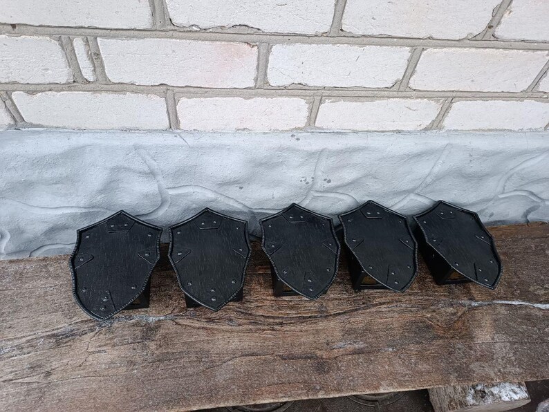 May include: Six black, shield-shaped decorative items with rivet details are arranged on a wooden surface. The shields have a textured appearance and are set against a backdrop of a brick wall and a gray surface.