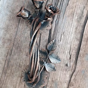 May include: A metal door handle shaped like a rose bush with three roses and leaves. The handle is a dark brown color with a copper finish.