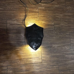 May include: A black, shield-shaped lamp with a textured surface, illuminated from within. The lamp is connected to a dark cord, casting a warm glow on the wooden floor. The design suggests a medieval or gothic theme.