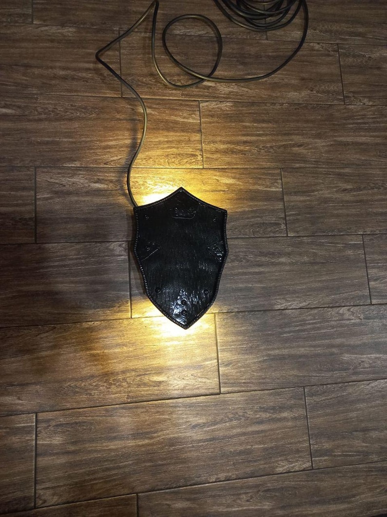 May include: A black, shield-shaped lamp with a textured surface, illuminated from within. The lamp is connected to a dark cord, casting a warm glow on the wooden floor. The design suggests a medieval or gothic theme.