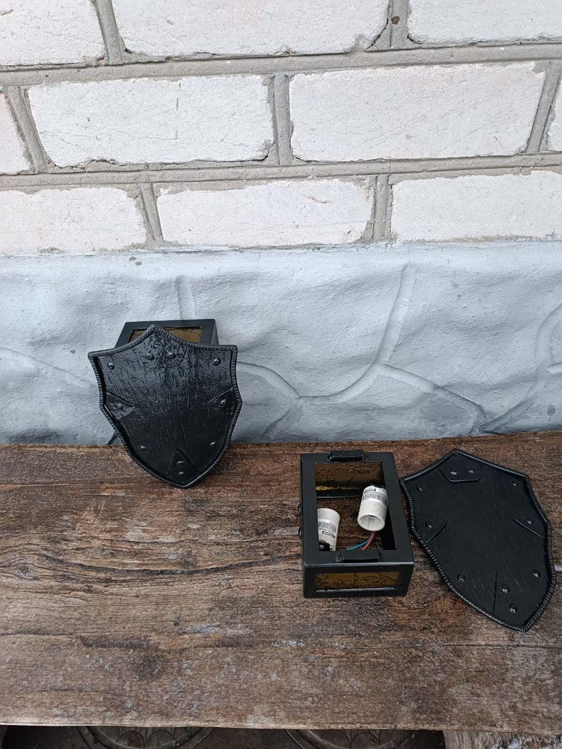 May include: Black, shield-shaped wall sconces with a textured finish. The sconces are made of metal and have a decorative design. The sconces are shown disassembled, with the electrical components visible. The sconces are on a wooden surface.