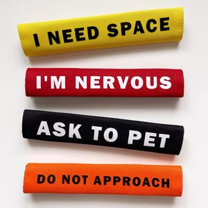 May include: Four rectangular dog accessories in yellow, red, black, and orange. Each has white text: "I NEED SPACE", "I'M NERVOUS", "ASK TO PET", and "DO NOT APPROACH". These are designed to communicate a dog's needs.