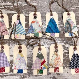May include: Ten rectangular cream-colored paper tags, each featuring a fabric Christmas tree appliqué. The trees are crafted from various patterned and colored fabric scraps, topped with a small button, and include a brown twine hanging loop.