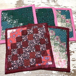 Puede incluir: Cuatro posavasos acolchados hechos a mano con bordes rosa y rojo. Los posavasos están hechos de tela de patchwork con patrones florales en tonos verdes, rojos y rosas.