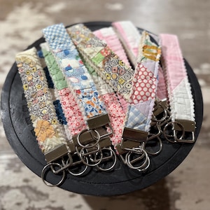 以下が含まれることがあります： 花柄、水玉模様、幾何学模様など、さまざまな模様のハンドメイド布製キーホルダーのコレクション。 各キーホルダーは金属製の留め金とキーリングを備え、パッチワークの美学を示しています。