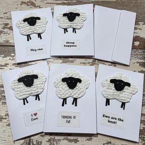 Op de afbeelding: Witte wenskaarten met een schapenontwerp. Elke kaart heeft een getextureerd wit schaap met een zwart gezicht en poten. Tekst op de kaarten is "Hey ewe", "sheep happens", "I ❤️ Ewe", "Thinking of Ewe" en "Ewe are the best!"