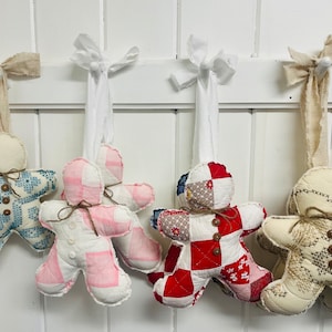 May include: Five handmade gingerbread decorations hanging from hooks on a white wall. The decorations are made from patchwork fabric in various colours and patterns, including blue, pink, red, and white. Each decoration has a brown twine bow and a button for a face.