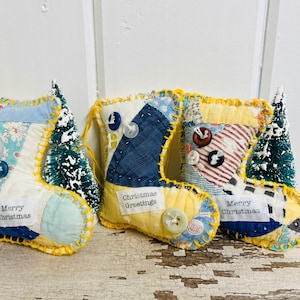 May include: Three handmade Christmas stocking ornaments made from patchwork fabric with yellow trim. Each stocking has a different message: "Merry Christmas", "Christmas Greetings", and "Merry Christmas".