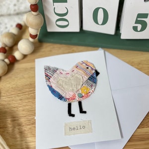 May include: A handmade greeting card with a patchwork bird design. The bird is made from fabric scraps in pink, blue, and yellow. The card includes the word "hello" on a small fabric tag. A white envelope is included with the card.