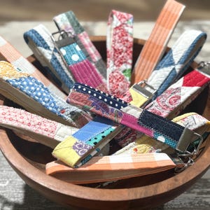 Op de afbeelding: Een verzameling kleurrijke stoffen sleutelhangers in een houten kom. De sleutelhangers hebben verschillende patronen, waaronder stippen, bloemenprints en patchwork-ontwerpen. Elke sleutelhanger heeft een metalen sluiting.