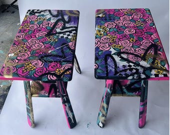 May include: Two small wooden stools with black, pink, and purple painted floral designs. The stools have a simple, four-legged design.