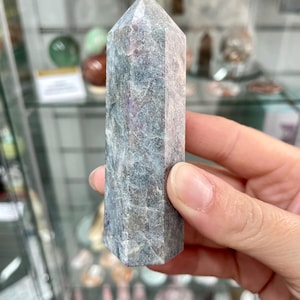 May include: A hand holds a tall, faceted crystal point. The crystal is a mix of grey, blue, and purple hues, with a textured surface and a pointed top. Other crystals and decorative items are visible in the background.