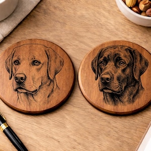 May include: Two round wooden coasters with engraved Labrador Retriever portraits. One shows a light brown dog, the other a dark brown dog. The coasters are displayed on a wooden surface.