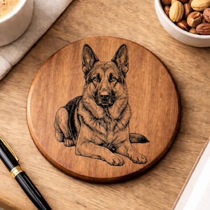 May include: A round wooden coaster with a detailed etched image of a German Shepherd dog. The dog is lying down, facing forward, with its ears perked up. The coaster is brown and sits on a wooden surface.