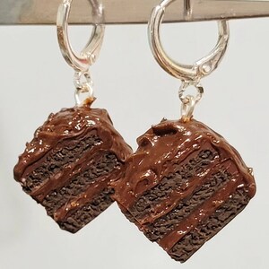 May include: A pair of silver hoop earrings with a chocolate cake charm hanging from each hoop. The cake charms are brown and have a chocolate frosting design.