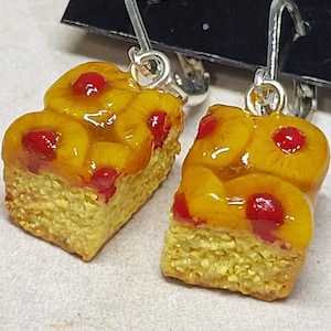 May include: A pair of silver dangle earrings featuring miniature yellow cake slices topped with red cherries and a clear glaze.