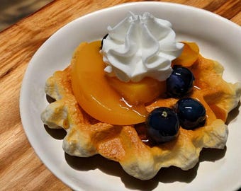 May include: A golden-brown waffle topped with a peach slice, whipped cream, and blueberries, served on a white plate. The waffle has a textured surface and is placed on a wooden table.