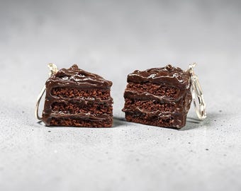 May include: A pair of earrings shaped like triangular slices of chocolate cake. Each piece features three distinct layers of dark brown cake with thick, glossy chocolate frosting on the top and between the layers, attached to silver-toned metal hooks.