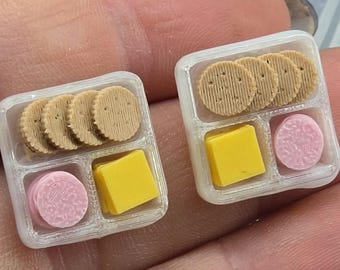 May include: Miniature food trays with crackers, cheese, and pink cookies. The crackers are round with a textured surface. The cheese is a yellow square, and the cookies are pink and round. The trays are white.