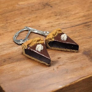 May include: A pair of chocolate pie slice earrings with a white dollop of whipped cream on top. The earrings are made of polymer clay and have silver hooks.