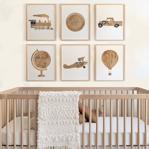 May include: A wooden crib with a white blanket and brown pillows. Six framed prints hang on the wall above the crib. The prints feature images of a train, compass, truck, globe, airplane, and hot air balloon.