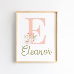 May include: Framed print featuring the letter "E" in a soft coral color, adorned with a floral arrangement. The name "Eleanor" is written in a cursive, olive green font below the letter. The artwork is set against a white background.