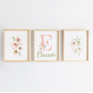 May include: Three framed floral art prints. The prints feature watercolor flowers in shades of pink and white, with green accents. One print displays the letter "E" with the name "Eleanor" below. The frames are light wood.