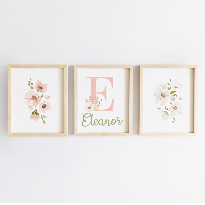 May include: Three framed floral art prints. The prints feature watercolor flowers in shades of pink and white, with green accents. One print displays the letter "E" with the name "Eleanor" below. The frames are light wood.