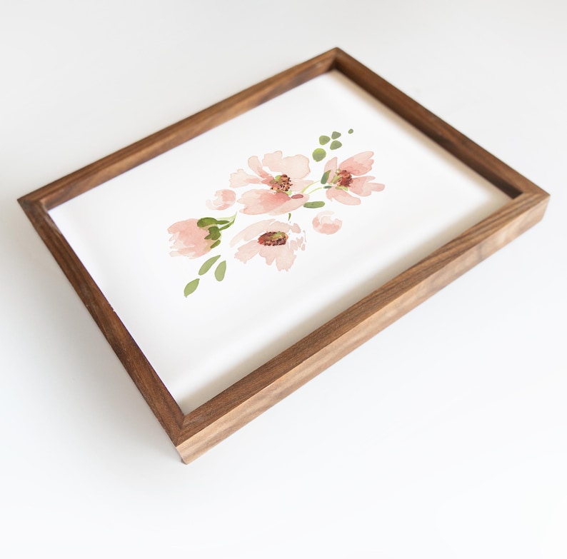 May include: A framed watercolor print featuring a cluster of pink flowers with green leaves. The artwork is set against a white background and encased in a rectangular, dark brown wooden frame. The floral design has a soft, delicate appearance.