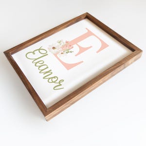 May include: Wooden framed art print featuring the name "Eleanor" in a green script font. A large pink letter "E" is decorated with a floral arrangement. The frame is made of dark brown wood.