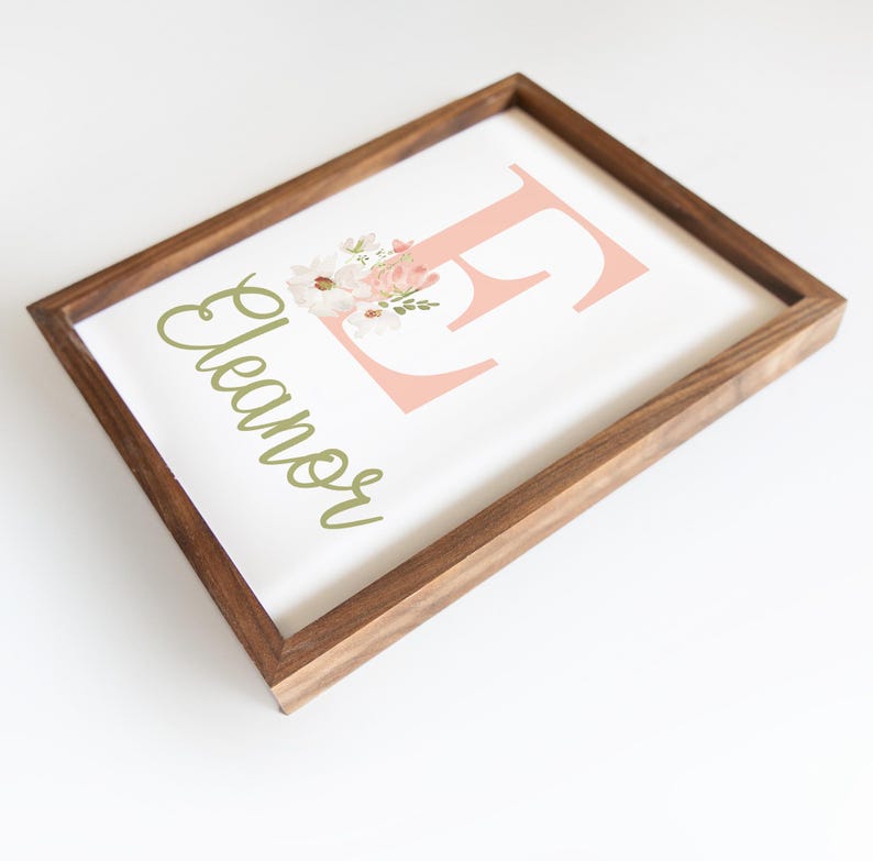 May include: Wooden framed art print featuring the name "Eleanor" in a green script font. A large pink letter "E" is decorated with a floral arrangement. The frame is made of dark brown wood.