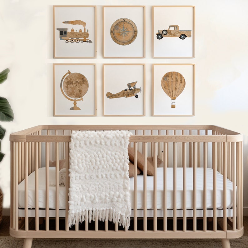 May include: A wooden crib with a white blanket and brown pillows. Six framed prints hang on the wall above the crib. The prints feature images of a train, compass, truck, globe, airplane, and hot air balloon.