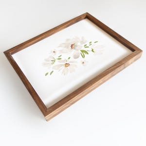 May include: A framed watercolor painting featuring white flowers with brown centers and green leaves. The artwork is set in a rectangular wooden frame with a dark brown finish. The painting is on a white background.