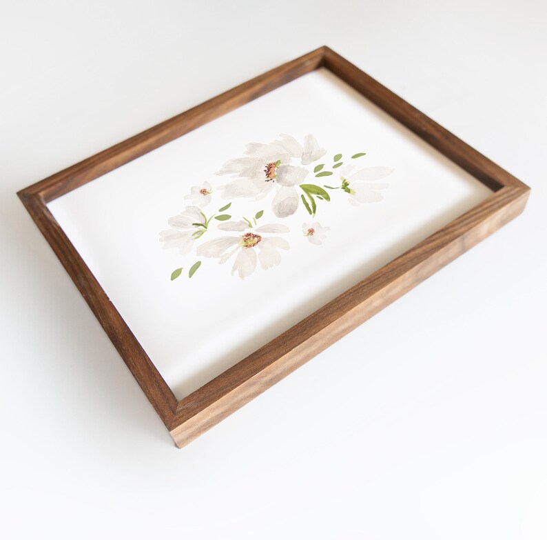 May include: A framed watercolor painting featuring white flowers with brown centers and green leaves. The artwork is set in a rectangular wooden frame with a dark brown finish. The painting is on a white background.