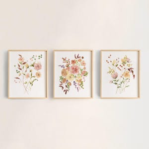 May include: Three rectangular wall art prints in light wood frames, each featuring a watercolor illustration of wildflowers and foliage in shades of pink, yellow, orange, and green against a white background, arranged in a horizontal row.