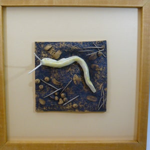 May include: A framed ceramic artwork depicting a white slug on a textured brown background with various small brown and white shapes.