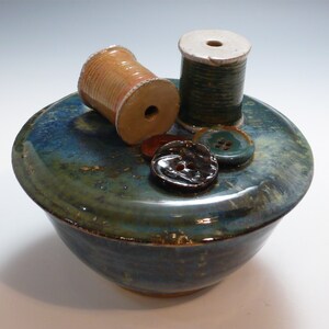 May include: A ceramic bowl with a blue and green glaze, decorated with two spools of thread, one brown and one green, and three buttons.