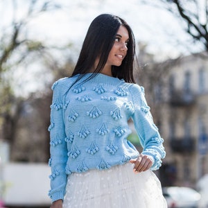 May include: A woman wearing a light blue knitted sweater with pom-poms and a white tulle skirt with white polka dots.